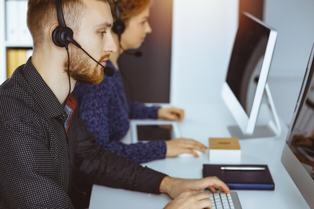 Empresário ruivo falando pelo fone de ouvido perto de sua colega enquanto está sentado no escritório ensolarado moderno. Grupo de pessoas diversas no call center.