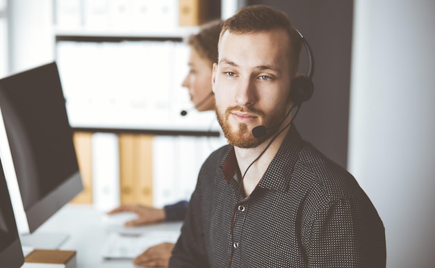 Empresário ruivo falando pelo fone de ouvido perto de sua colega enquanto está sentado no escritório moderno. Grupo de pessoas diversas no call center. Telemarketing e atendimento ao cliente.