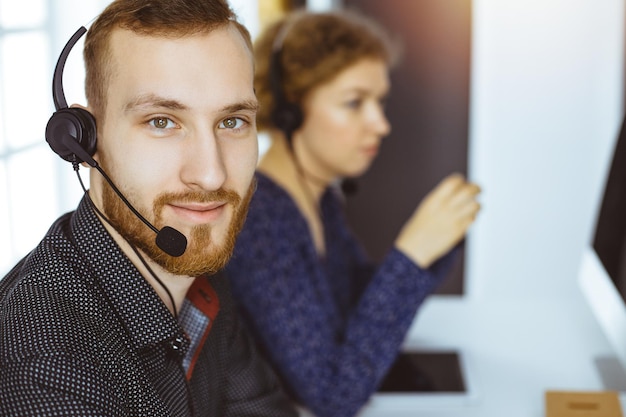 Empresário ruivo falando por fone de ouvido perto de sua colega enquanto está sentado no escritório ensolarado moderno Grupo de pessoas diversas em call center