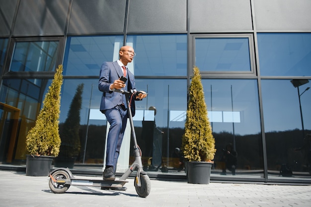 Empresario con scooter eléctrico de pie delante del edificio de negocios moderno hablando por teléfono.