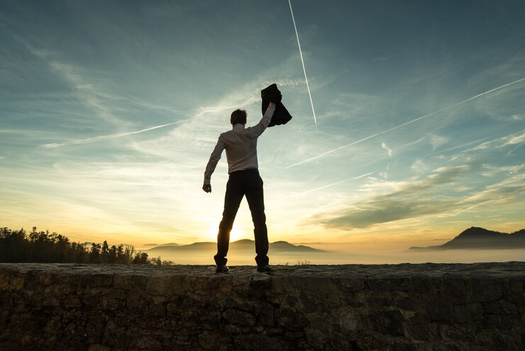 Foto empresário segurando sua jaqueta ao pôr do sol enquanto ele fica em uma parede com vista para uma cena de montanha enevoada silhueta contra o céu colorido brilhante com rastos.