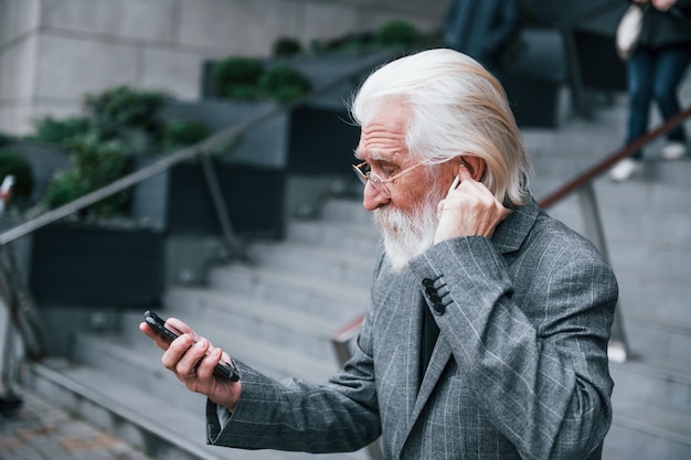 Empresário sênior em roupas formais com cabelos grisalhos e barba está ao ar livre tentando fones de ouvido sem fio com telefone