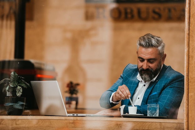 Empresario senior moderno sentado en un café y usando una computadora portátil y un teléfono inteligente