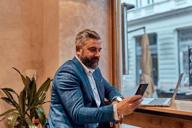 Empresario senior moderno sentado en un café y usando una computadora portátil y un teléfono inteligente.