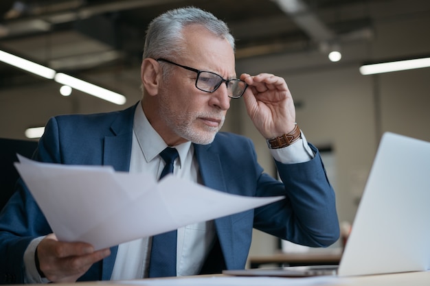 Empresario senior pensativo con contrato de lectura portátil, buscando en línea