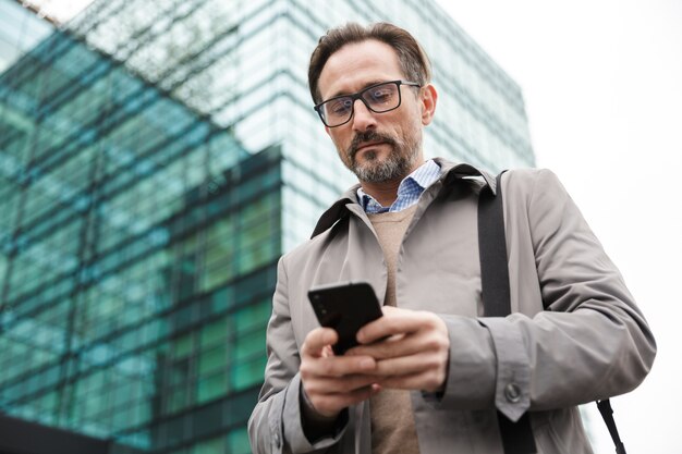 empresário sério bonito usando óculos usando o celular em pé perto de um prédio de vidro na área urbana