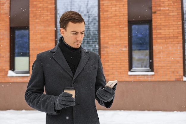 Empresário sério em casaco de inverno surfando smartphone e tomando café na rua da cidade