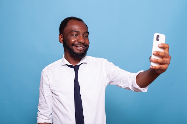 Empresario sonriente divirtiéndose tomando una foto selfie usando la cámara frontal de un teléfono inteligente sobre fondo azul. Feliz trabajador independiente sosteniendo un teléfono inteligente en una videoconferencia posando informal.