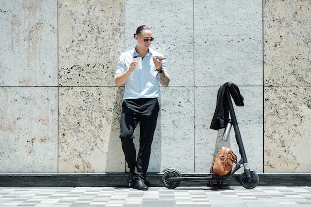 Empresario sonriente con gafas de sol bebiendo café para llevar y grabando un mensaje de voz para el cliente o compañero de trabajo