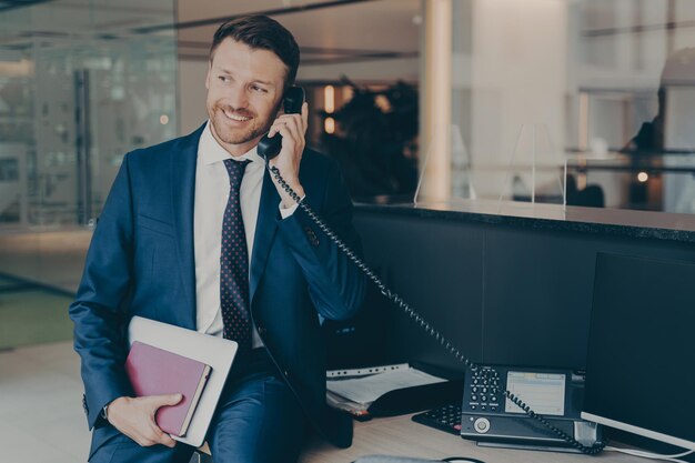 Empresario sonriente hablando por teléfono sentado en la oficina en la parte superior del escritorio sosteniendo el portátil y la agenda