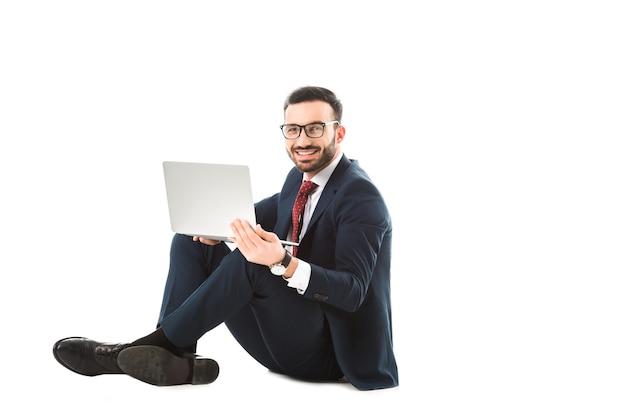 Empresario sonriente mirando a la cámara mientras está sentado y usando una computadora portátil con fondo blanco