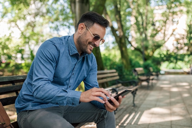 Foto empresario sonriente en red en un teléfono inteligente mientras está sentado en un banco en el parque