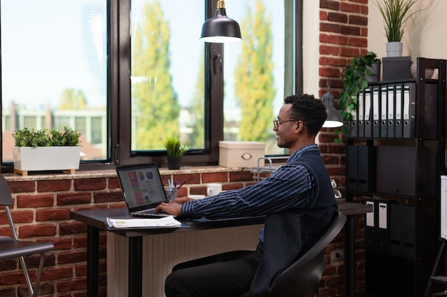 Empresario sonriente sentado en el escritorio en la oficina de inicio usando una computadora portátil para analizar los resultados comerciales. Empresario casual mirando la pantalla de la computadora portátil trabajando en gráficos de estadísticas de ventas.
