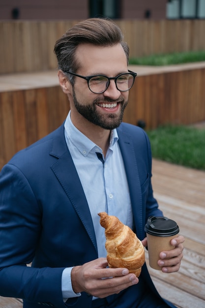 Empresario sonriente sosteniendo una taza de café comiendo croissant en la calle a la hora del almuerzo pausa para el café