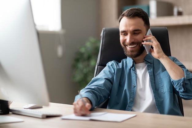 Empresario sonriente tomando notas mientras tiene una conversación telefónica en la oficina