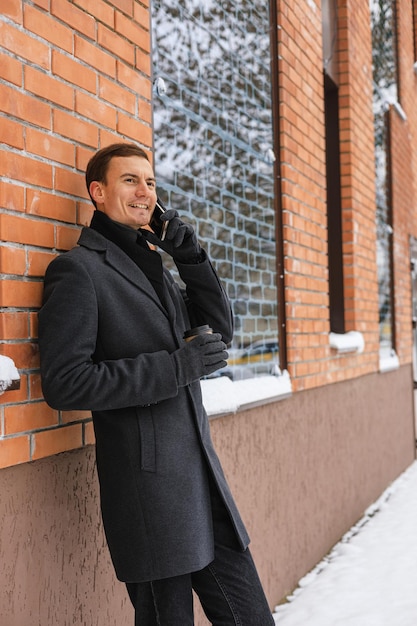 Empresário sorridente com casaco de inverno com uma xícara de café fazendo chamada para smartphone na rua da cidade