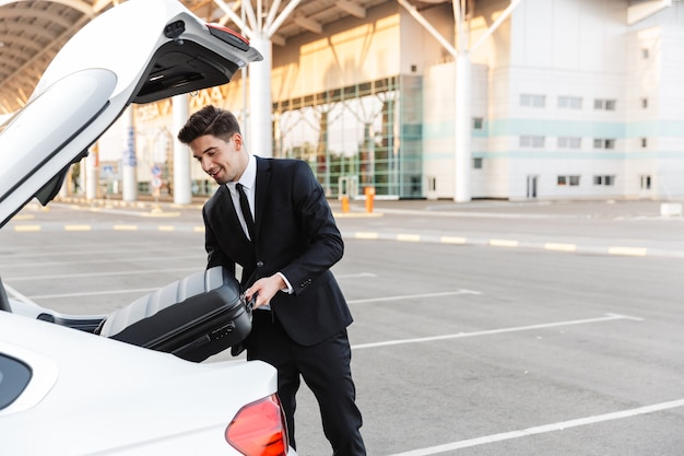 empresário sorridente com roupa formal colocando a mala no porta-malas do carro no estacionamento perto do aeroporto ao ar livre