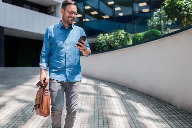 Empresário sorridente enviando mensagens no celular enquanto caminhava contra o prédio de escritórios