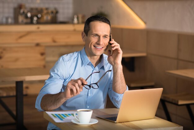 Empresário sorridente falando no smartphone e segurando óculos no café