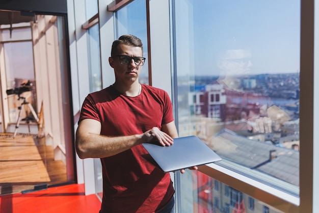 Empresário sorridente fica ao lado da janela enquanto trabalha em um laptop conceito de multitarefa um freelancer ocupado em óculos segura um laptop nas mãos enquanto está perto da janela smart casual