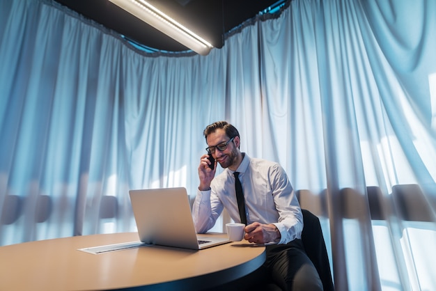 Empresário sorridente pegando o telefone e bebendo café enquanto está sentado na sala de conferências. Na frente dele, o laptop na mesa. Na cortina de fundo.