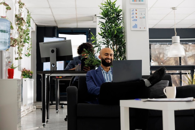 Empresário sorridente sentado no sofá no escritório enquanto faz uma videochamada com o cliente. Gerente executivo discutindo a estratégia do projeto com trabalhador remoto em videoconferência no laptop