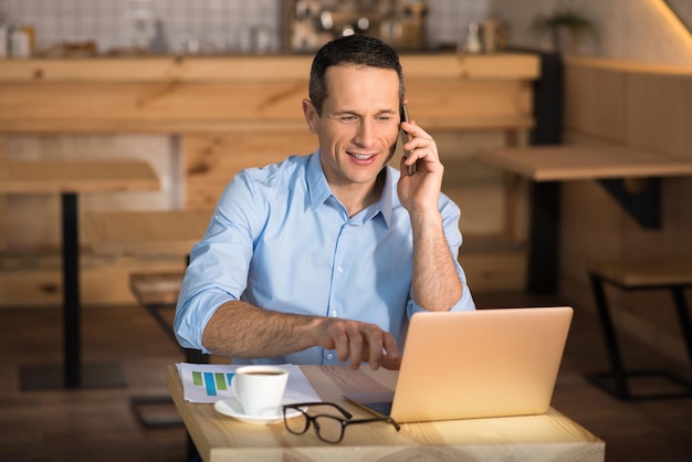 Empresário sorridente trabalhando no laptop no café enquanto fala no smartphone