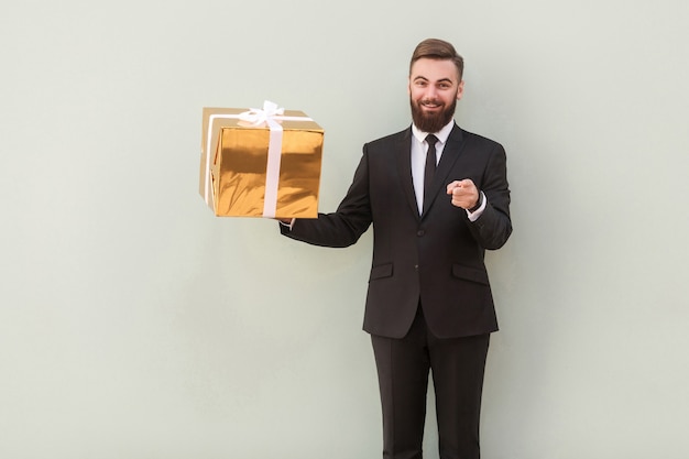 Empresario sosteniendo caja de regalo, señalando con el dedo a la cámara y sonriendo. Tiro de interior