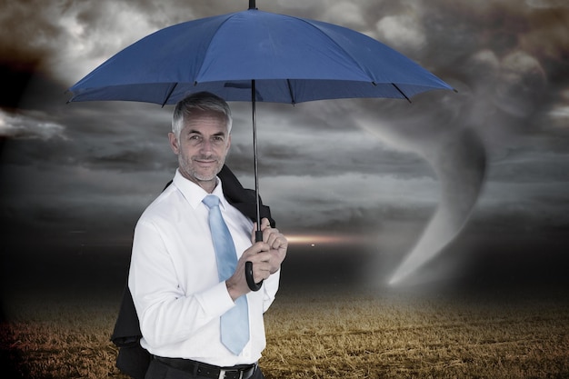Empresario sosteniendo paraguas azul contra el cielo tormentoso con tornado sobre campo