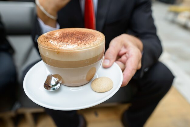 Empresario sosteniendo una taza de café capuchino con chispas de chocolate en espuma y galletas en el lateral