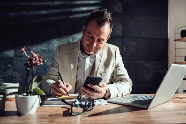 Empresario sosteniendo teléfono inteligente y escribiendo en una notas adhesivas