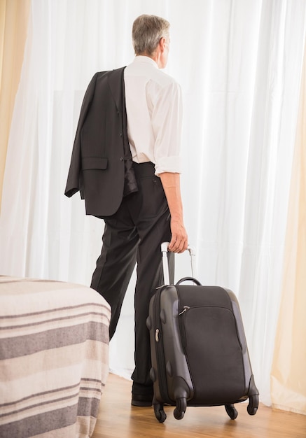 Empresario con su maleta en la habitación del hotel mirando la ventana Vista posterior