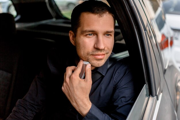 Empresario en un taxi tiene un teléfono inteligente y mira por la ventana. Paseos de pasajeros en el retrato de primer plano del asiento trasero