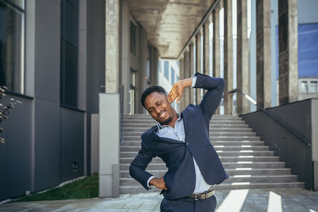 Empresário tem fortes dores nas costas no trabalho, homem afro-americano freelancer perto do escritório segurando as costas deprimido tentando fazer massagem