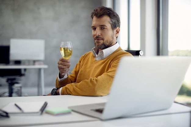 Empresario tomando una copa de vino mientras sale en línea con una laptop en la oficina