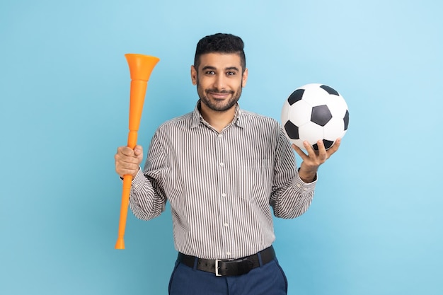 Empresário torcendo pelo time de futebol favorito segurando chifre e bola nas mãos olhando para a câmera