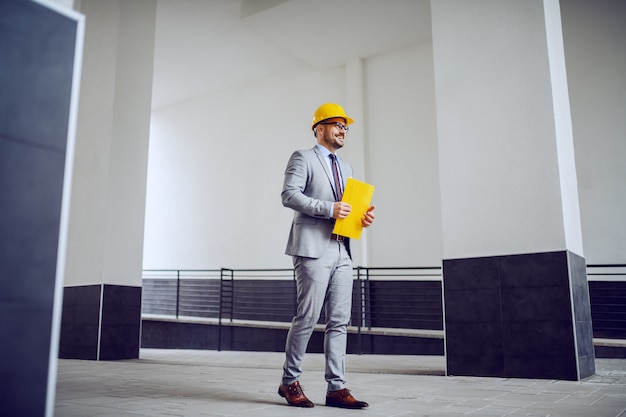 Empresario en un traje gris con un casco protector amarillo en la cabeza caminando al aire libre y con un archivo amarillo en sus manos