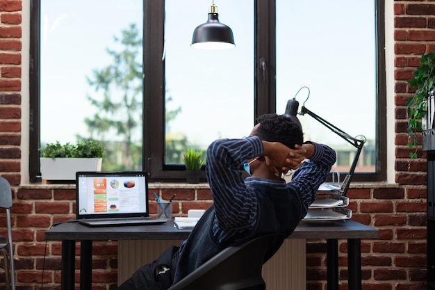 Empresário usando máscara de proteção covid que se estende no final do árduo dia de trabalho na cadeira do escritório na mesa. Proprietário de empresa de inicialização relaxante olhando para laptop com indicadores de desempenho.