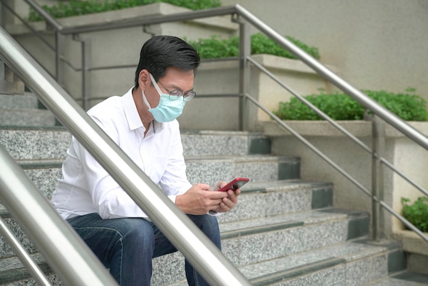 Empresário usando máscara facial sentado na escada usando e lendo seu telefone