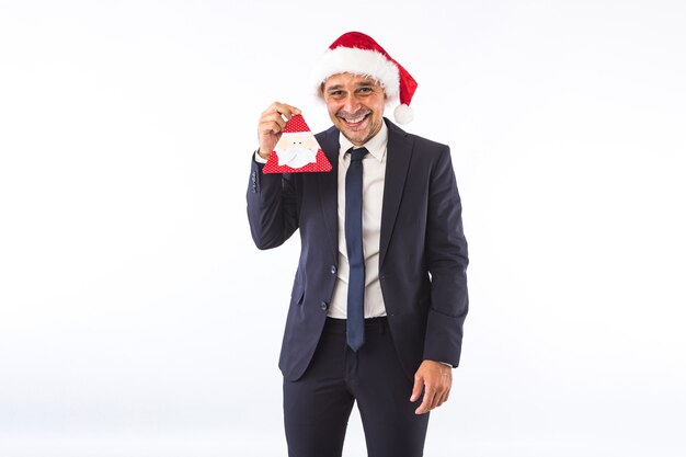 Empresário vestido de terno, gravata e chapéu de Natal do Papai Noel, sorrindo, segurando um enfeite de Papai Noel, sobre fundo branco. Conceito de celebração de Natal.