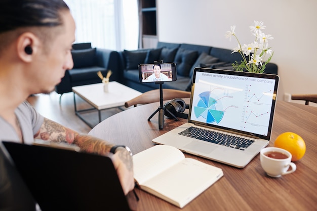 Empresario con videollamada con un colega vietnamita y discutiendo el diagrama en la pantalla del portátil cuando se trabaja desde casa