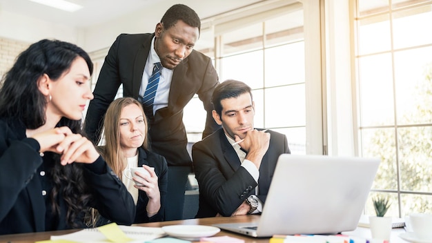 Empresarios africanos de pie en la parte de atrás de colegas caucásicos mirando en la computadora portátil en una reunión interna del grupo empresarial. Diversidad de cooperación de empresarios.