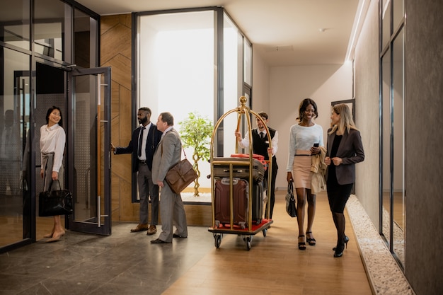 Foto empresários contemporâneos se comunicando enquanto o carregador se move entre eles e empurra o carrinho com a bagagem dentro do hotel