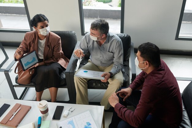 Empresários em máscaras médicas discutindo gráficos e diagramas com dados financeiros na reunião