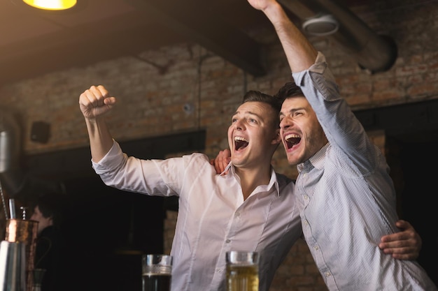 Empresarios de éxito bebiendo cerveza y gritando mientras ven la televisión juntos en el bar. Hombres seguros divirtiéndose