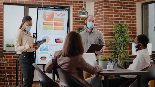 Empresários explicando o projeto financeiro e a apresentação aos colegas na sala de reuniões. Colegas de trabalho apresentando análise de gráficos de dados no monitor na reunião durante a pandemia de coronavírus.