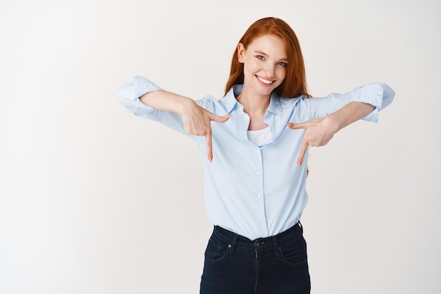 Empresários Jovem ruiva bonita apontando os dedos para baixo mostrando o logotipo da empresa e sorrindo em pé sobre fundo branco