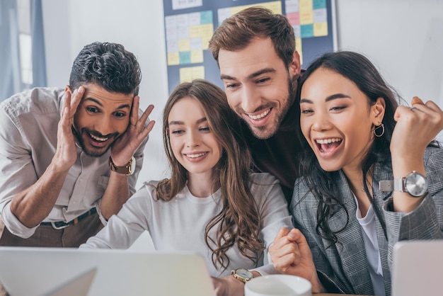 Empresarios multiculturales alegres y empresarias mirando portátil en la oficina