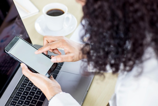 Los empresarios sentados en la oficina sobre el escritorio de trabajo con computadora y gráficos y café, sostienen teléfonos inteligentes con pantallas en blanco. Tomado desde el ángulo de visión superior