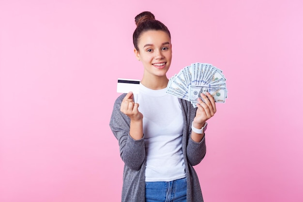 Empréstimo bancário. Retrato de adolescente morena alegre com penteado de coque em roupas casuais, mostrando notas de dólar e cartão de crédito, sorrindo para a câmera. tiro de estúdio interior isolado no fundo rosa